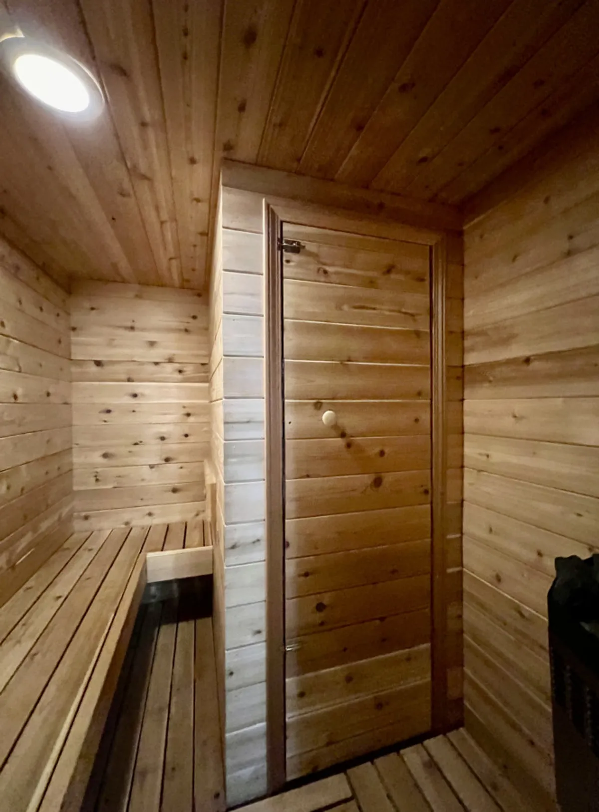 A close-up interior view of a sauna with wooden benches, a wooden door, and a heating unit. The warm wood tones and intimate layout highlight the space’s cozy feel and focus on self-care.