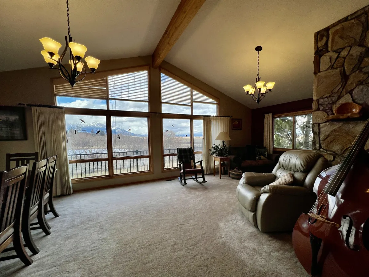 This inviting interior combines a comfortable seating area, stone fireplace, and a dining table, all oriented towards expansive windows showcasing the outdoors. The layout encourages easy conversation and togetherness, with cozy furnishings that enhance the sense of warmth and community.