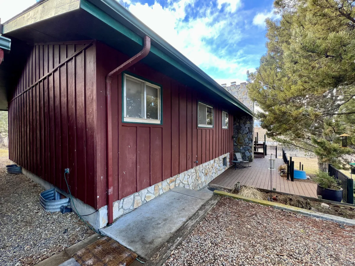 A wooden deck with a stone wall and red wooden siding nestled among mature trees, creating a cozy and welcoming outdoor space. There are signs of outdoor furniture and space for gatherings or grilling, making it ideal for social events.