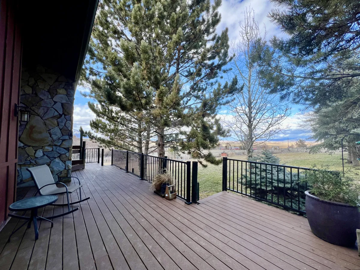 A spacious wooden deck wraps around the house, bordered by tall pine trees and overlooking a lush grassy area. The scene captures the deck’s expansive size, ideal for hosting gatherings or relaxing outdoors, with distant mountain landscape visible in the background.