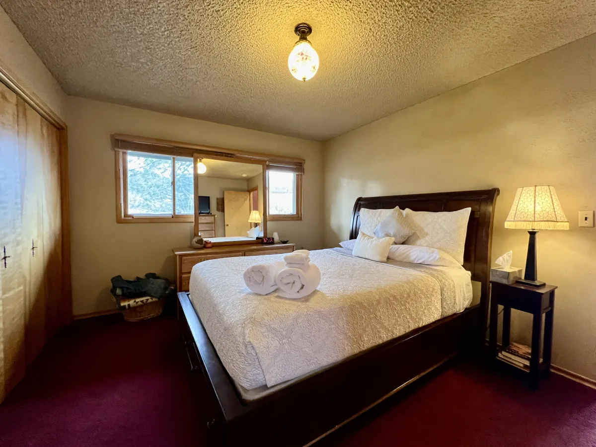 A warm, inviting bedroom featuring a queen-sized bed with plush linens, a mirror, a nightstand with a glowing lamp, and fresh towels placed thoughtfully nearby. Natural light streams in through the window, enhancing the tranquil ambiance.
