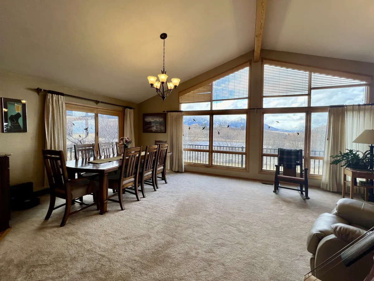A spacious dining area with a large table sits in front of floor-to-ceiling windows, allowing abundant natural light to pour in and offering sweeping views of the mountains beyond. A chandelier hangs above the table, setting a welcoming mood for gatherings. The scene is set for socializing and celebrating, with the landscape as a striking backdrop.