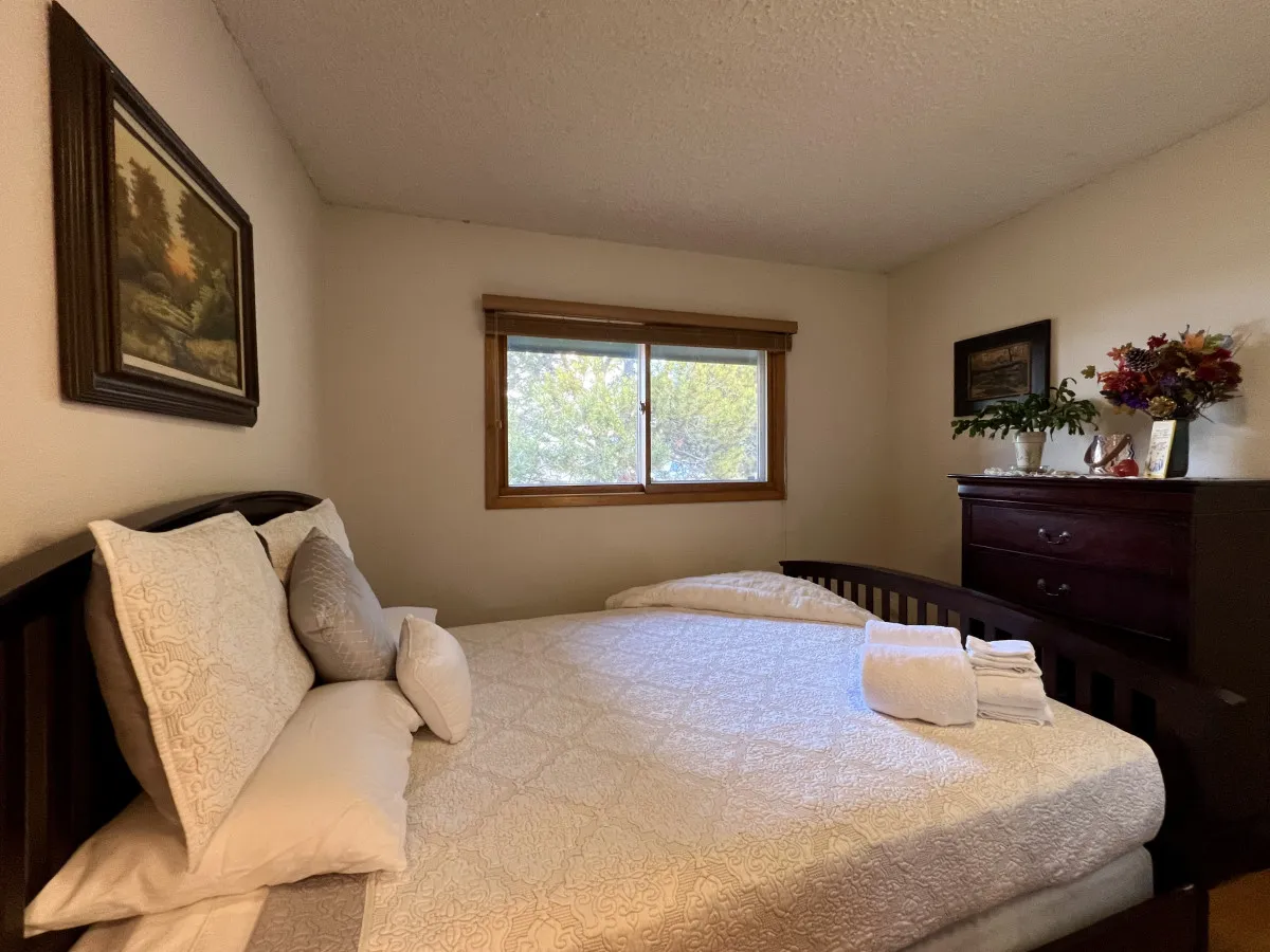 A tranquil bedroom featuring a bed with decorative pillows, framed artwork, a window spilling natural light, and a dresser adorned with plants. The soft palette and homey details evoke a sense of peaceful retreat.