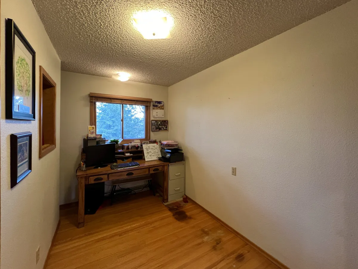 A dedicated office nook with a wooden desk, computer setup, and window offering an inspiring outdoor view. Framed artwork and organized surroundings create an atmosphere suited for focused work or quiet reflection.