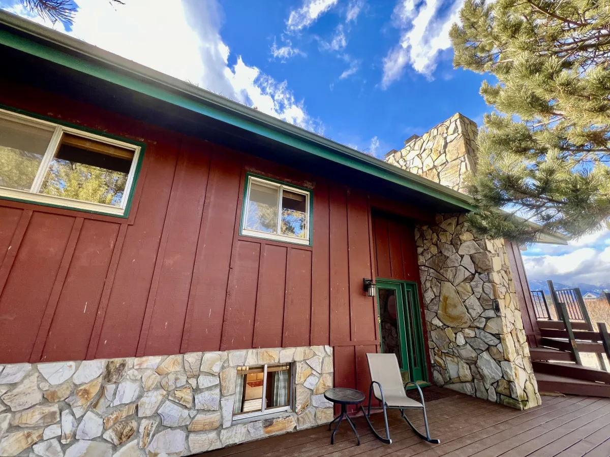 A cozy exterior shot of Elkhorn Views Home, featuring brown wooden siding, a stone wall, green trim, a deck area, and two comfortable chairs. The setting invites relaxation with views toward the surrounding landscape, perfect for enjoying sunsets or stargazing after a productive day.