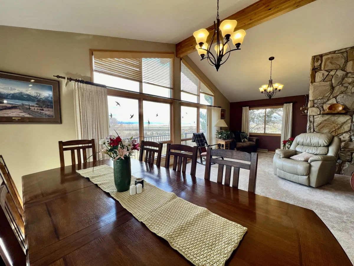The image showcases a sunlit, open-plan living and dining area with a generous dining table set before expansive floor-to-ceiling windows. These windows offer sweeping views of the Elkhorn Mountains and Baker Valley. A stone fireplace anchors the cozy seating area, inviting guests to gather and unwind while admiring the natural beauty beyond.
