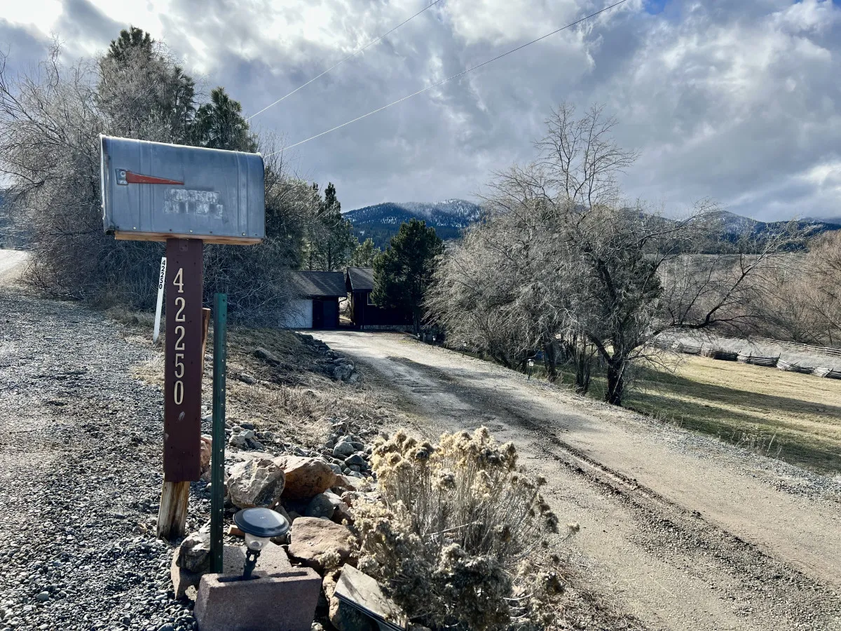 A serene scene showing a mailbox with the number 42250, a gravel driveway lined with trees, and a stunning mountain backdrop. The calm surroundings and open fields evoke a sense of peace and provide context for the home's scenic location.